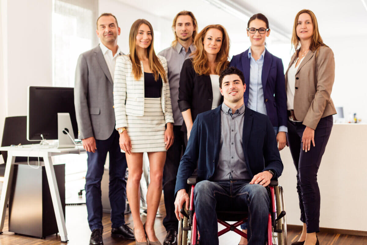 small business team posing for a group shot in their office. one man is sitting in a wheelchair.