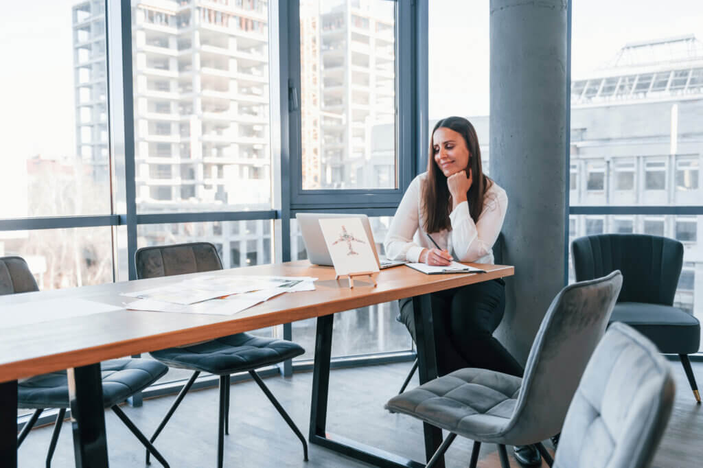 Sits by the table. Young woman in white formal clothes is indoors in the modern office.