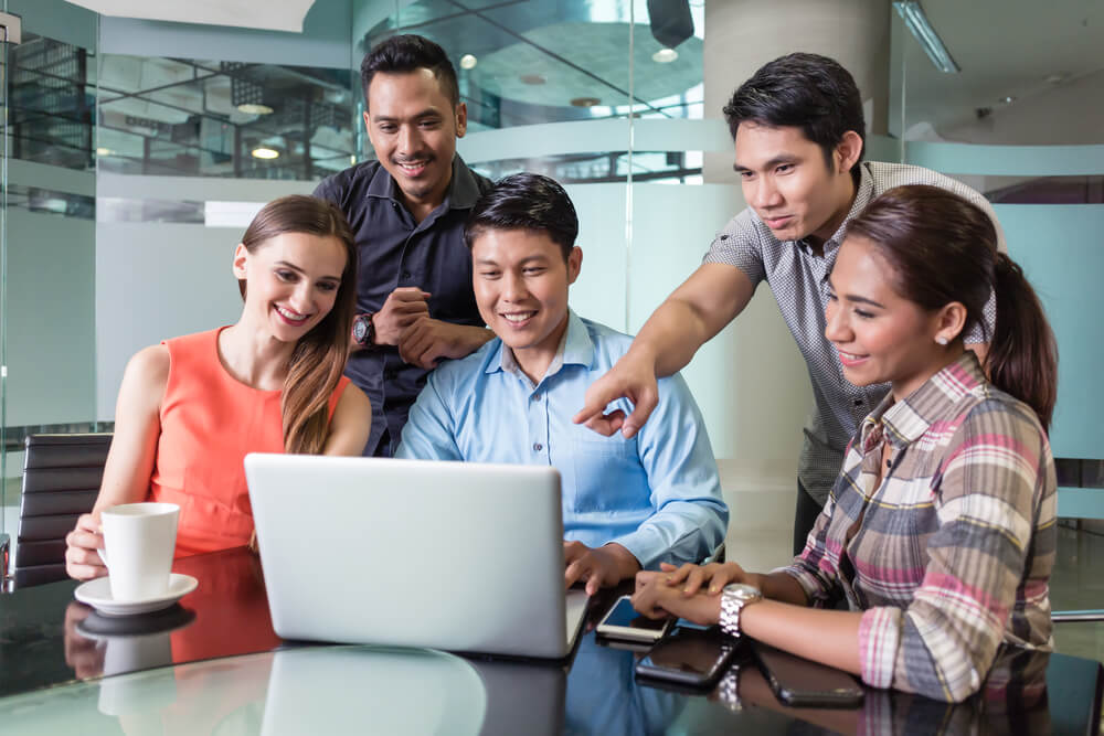 Multi-ethnic team of five cheerful employees smiling while watching a funny video or presentation on laptop in the meeting room of a multinational company